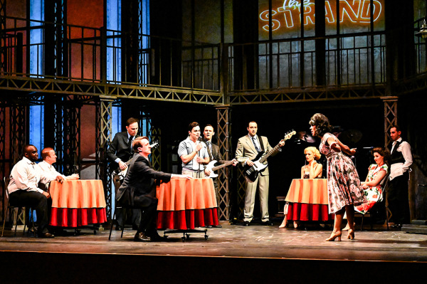 Actors on stage at a pub, sitting around tables while four actors play guitar and sing.